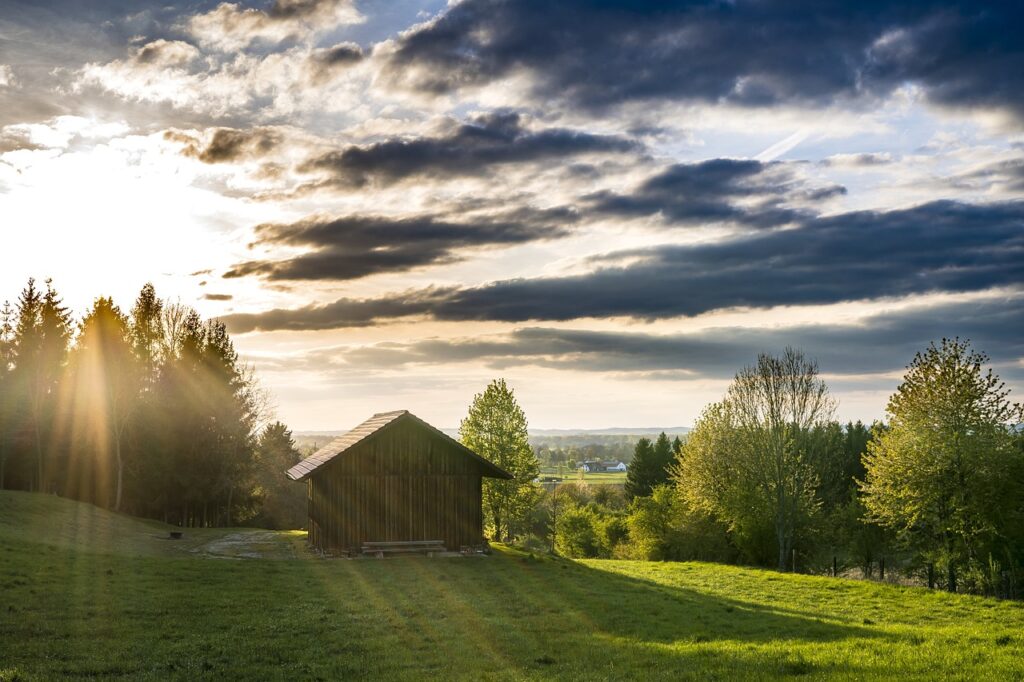 Eine hölzerne Scheune steht auf einer Wiese bei Sonnenuntergang, mit Bäumen und Sonnenstrahlen, die durch die Wolken scheinen.