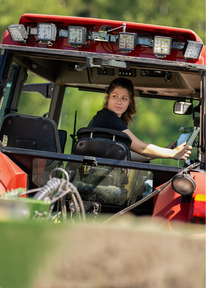 Eine Frau, die einen roten Traktor fährt, blickt über ihre Schulter zurück, während draußen grüne Bäume zu sehen sind.