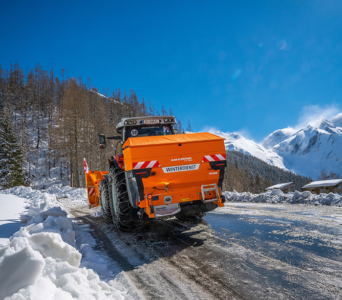 Ein orangefarbener Schneepflug räumt eine verschneite Bergstraße unter einem strahlend blauen Himmel mit schneebedeckten Gipfeln.