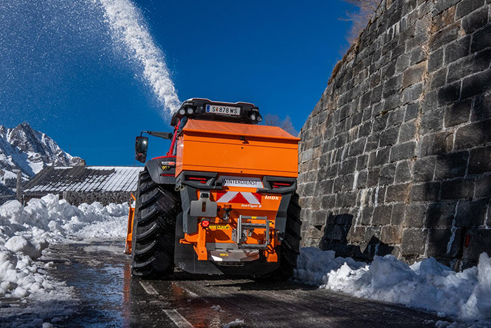 Orangefarbener Schneepflug, der an einem sonnigen Wintertag in der Nähe einer Steinmauer Schnee räumt, mit Bergen im Hintergrund.