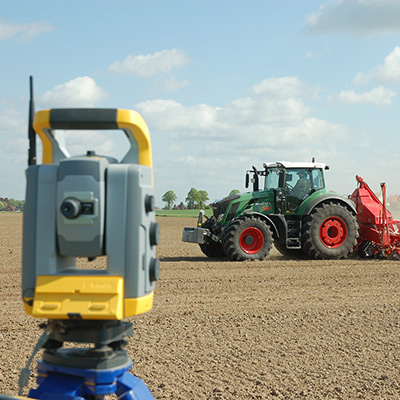 Ein Traktor arbeitet auf einem gepflügten Feld mit einem Vermessungsinstrument im Vordergrund unter blauem Himmel.