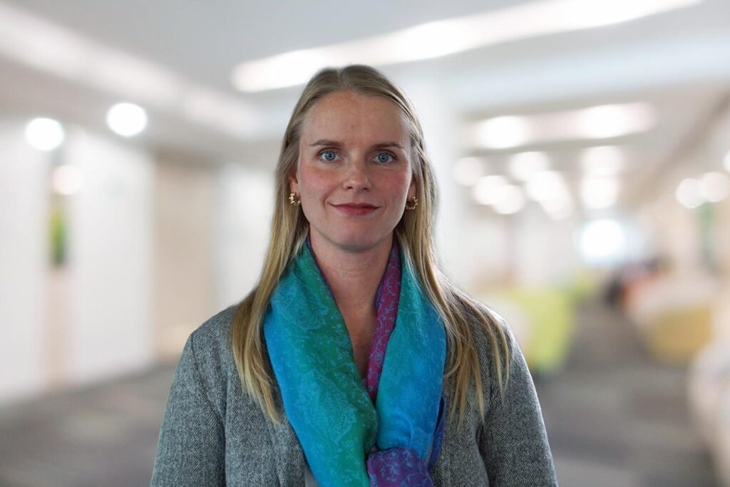 Eine Frau mit langen blonden Haaren und einem blauen Schal steht in einem verschwommenen Hausflur.