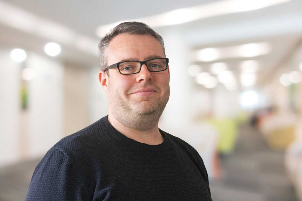 Mann mit Brille und schwarzem Pullover lächelt in einem hell erleuchteten, unscharfen Innenraum.