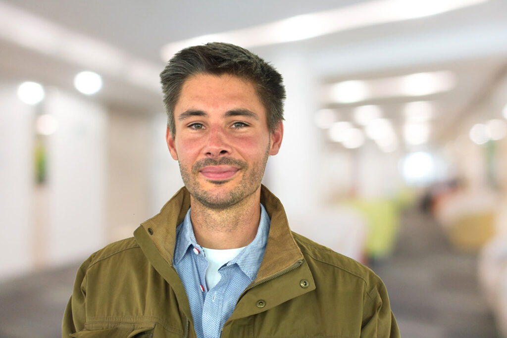 Ein Mann mit kurzen dunklen Haaren und Bart, der eine braune Jacke trägt, steht in einem hell erleuchteten Hausflur.