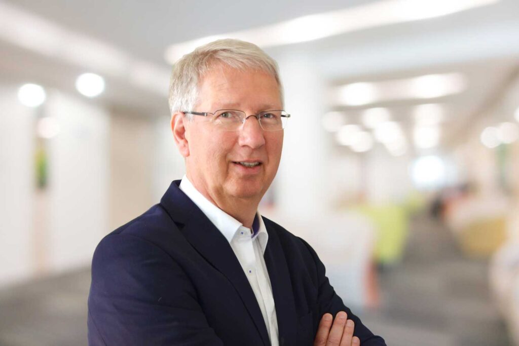 Ein lächelnder älterer Mann mit Brille in einer Anzugsjacke steht in einem hellen, unscharfen Innenraum.