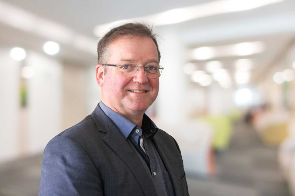 Ein Mann im Anzug und mit Brille lächelt in einer hell erleuchteten, verschwommenen Büroumgebung.