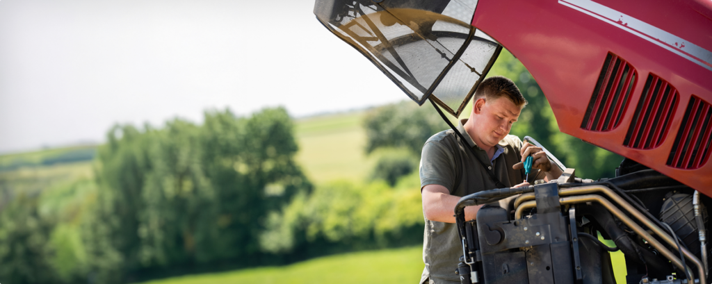Ein Mann repariert einen roten Traktormotor im Freien mit grünen Feldern und Bäumen im Hintergrund.