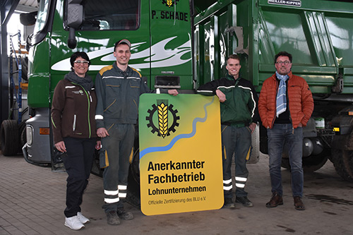 Vier Personen stehen lächelnd neben Lastwagen und halten ein Schild mit der Aufschrift Anerkannter Fachbetrieb Lohnunternehmen.