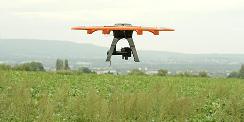 Orange-schwarze Drohne, die über ein grünes Feld mit in der Ferne sichtbaren Hügeln fliegt.