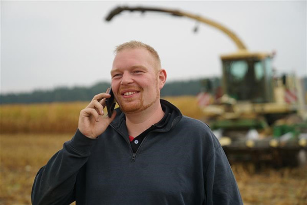 Ein lächelnder Mann auf einem Feld spricht mit einem Mobiltelefon, während im Hintergrund Landmaschinen zu sehen sind.