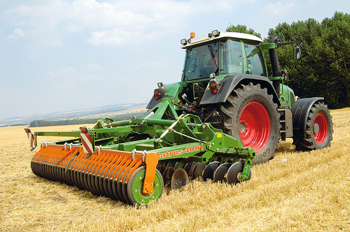 Ein Traktor mit einer grünen Scheibenegge arbeitet auf einem abgeernteten Feld unter einem teilweise bewölkten Himmel.