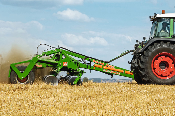 Ein grüner Grubber, der an einen Traktor angehängt ist, pflügt durch ein goldenes Feld unter einem blauen Himmel mit Wolken.