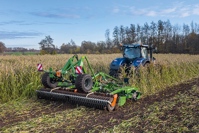 Ein blauer Traktor mit einem grünen Gerät pflügt unter einem teilweise bewölkten Himmel hohes Gras auf einem Feld.
