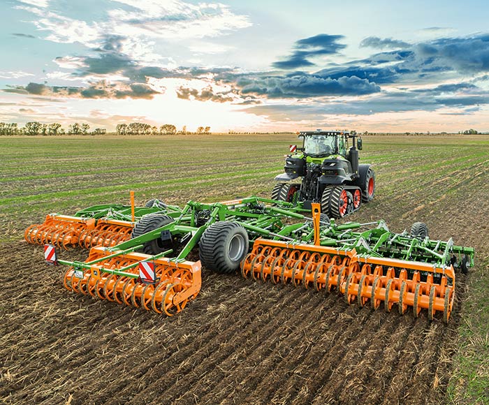 Ein großer Traktor zieht bei Sonnenuntergang einen grün-orangefarbenen Grubber über ein Feld, um den Boden zu bearbeiten.
