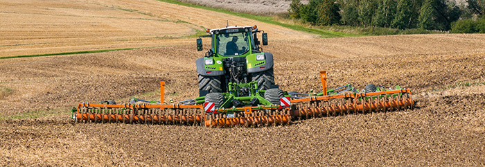 Ein grüner Traktor, der einen großen orangefarbenen Grubber zieht, pflügt an einem sonnigen Tag ein braunes Feld.