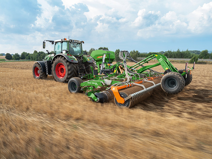 Ein grüner Traktor mit großen landwirtschaftlichen Geräten bewirtschaftet ein braunes Feld unter einem bewölkten Himmel.