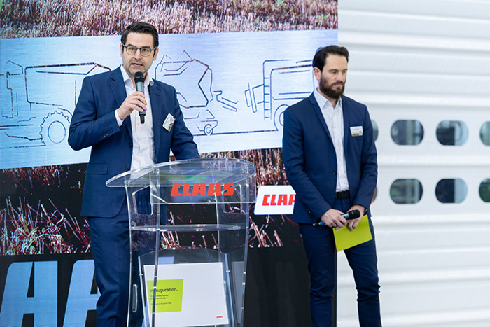 Zwei Männer in Anzügen sprechen auf einem Podium mit CLAAS Schriftzug, das vor einer Grafik mit Landmaschinen steht.