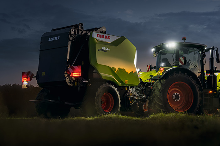 Ein Claas-Traktor mit angehängter Ballenpresse wird in der Dämmerung auf einem Feld mit leuchtenden Lichtern beleuchtet.