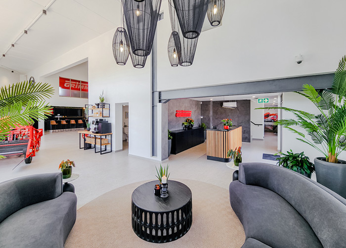 Moderne Büro-Lobby mit grauen Sofas, runder schwarzer Tisch, hängende Lichter, und grüne Pflanzen. Empfangsschalter im Hintergrund.
