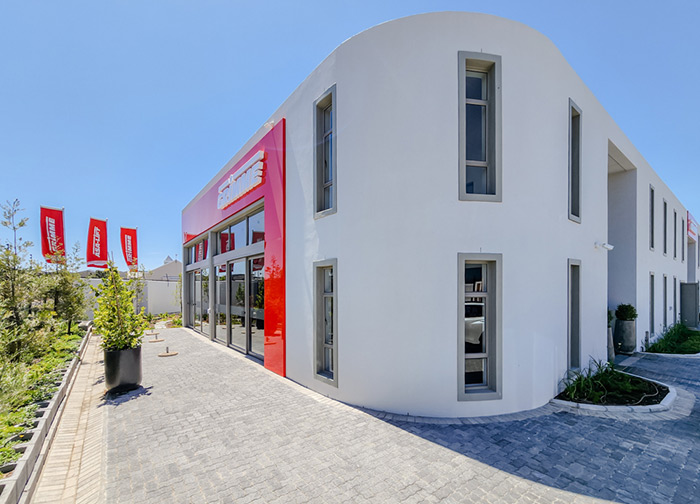 Modernes weißes Gebäude mit großen Fenstern, roten Akzenten und Flaggen, an einem sonnigen Tag mit blauem Himmel.