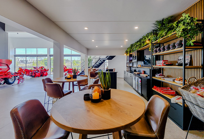 Moderne Showroom-Lounge mit runden Holztischen, Lederstühlen, Pflanzen und roten Motorrädern im Hintergrund.