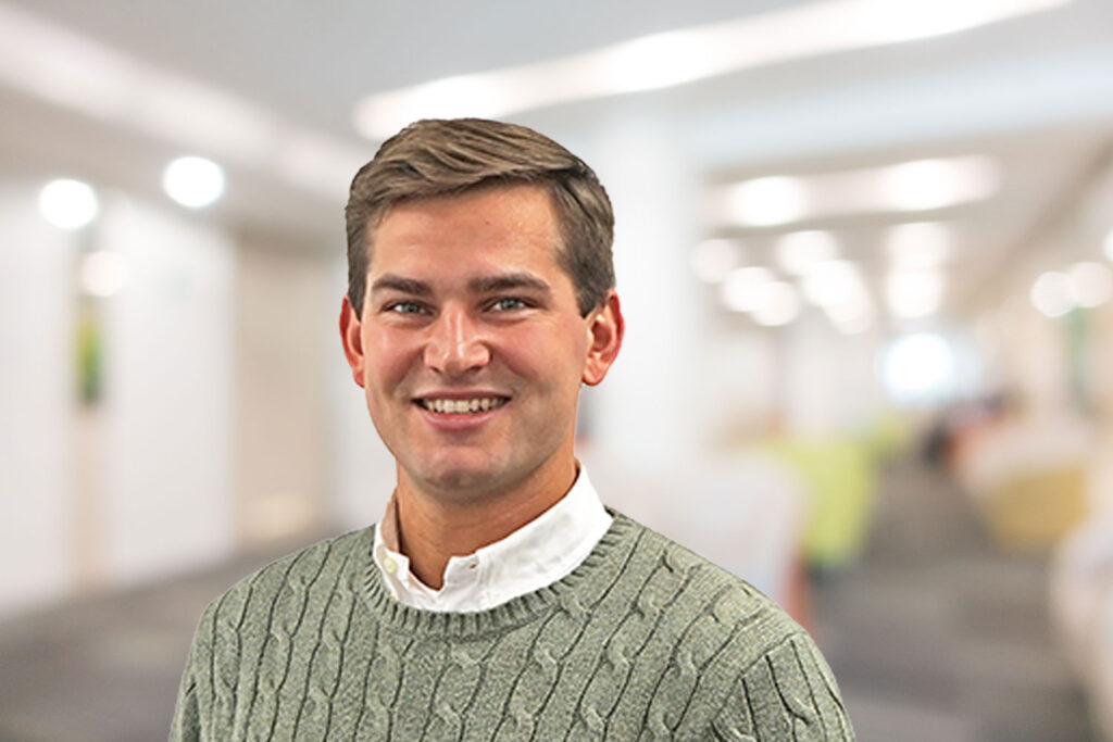 Ein lächelnder Mann in einem grünen Pullover steht in einem hell erleuchteten, verschwommenen Büroflur.