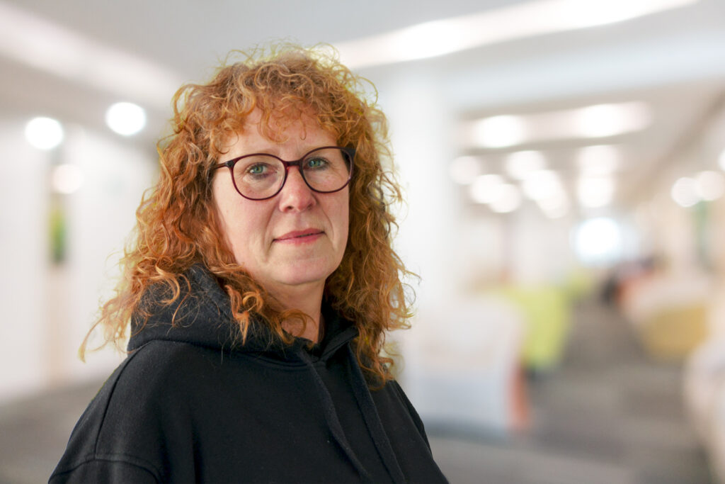 Frau mit roten Locken und Brille, schwarzer Kapuzenpulli, steht in einer unscharfen Innenraumumgebung.