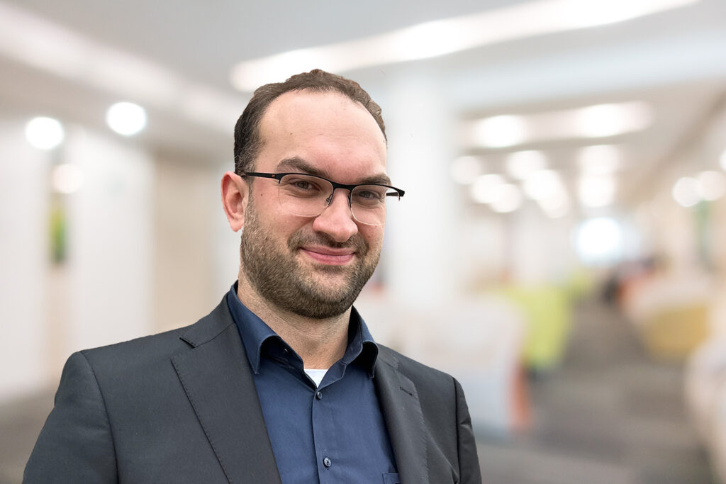 Ein Mann mit Brille und Anzug lächelt in einem hell erleuchteten, modernen Büroflur.