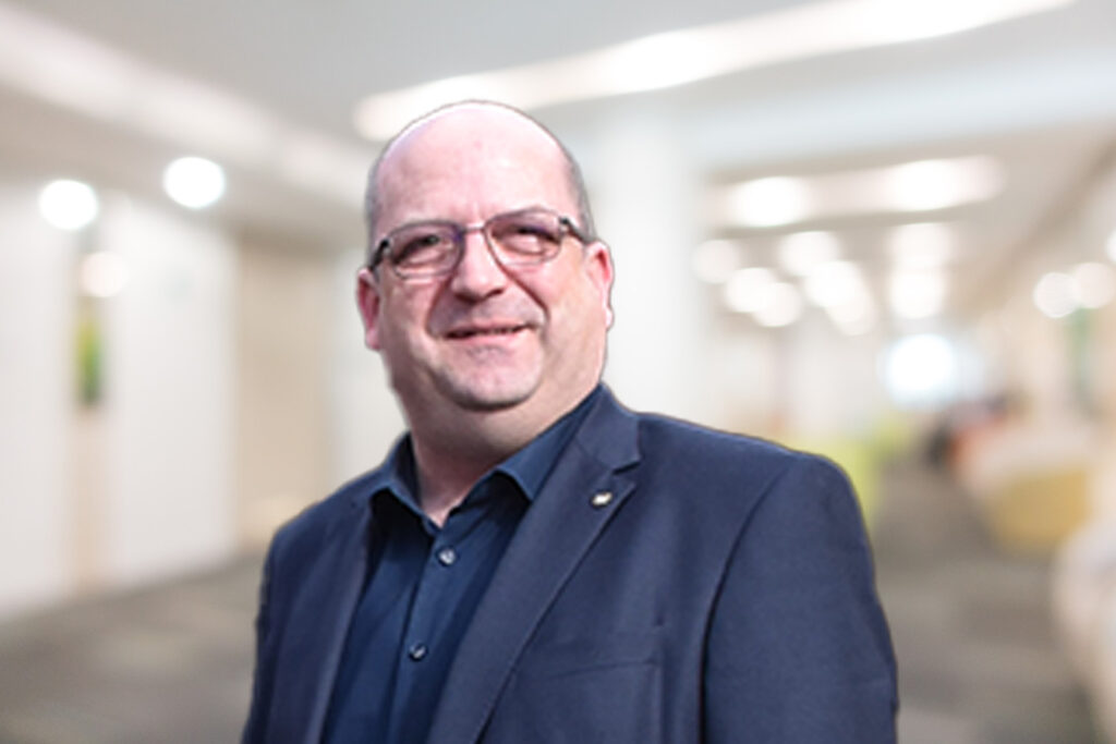 Ein Mann in einem dunklen Anzug und mit Brille lächelt in einem hellen, verschwommenen Flur.