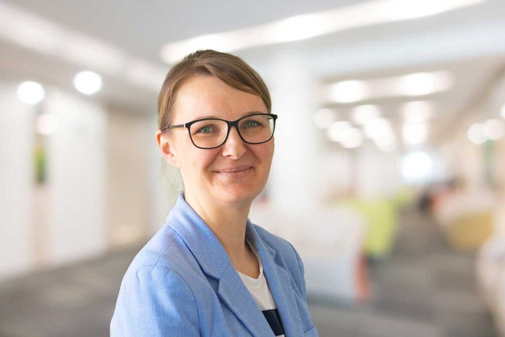 Frau mit Brille und blauem Blazer lächelt in einer hellen, verschwommenen Büroumgebung.