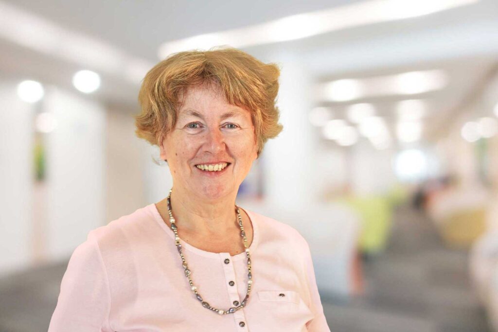 Lächelnde ältere Frau mit kurzen Haaren in einem rosa Oberteil und einer Halskette, die in einem hellen, verschwommenen Flur steht.