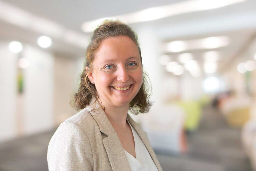 Frau mit lockigem Haar in einem beigen Blazer lächelt in einer hellen, verschwommenen Büroumgebung.