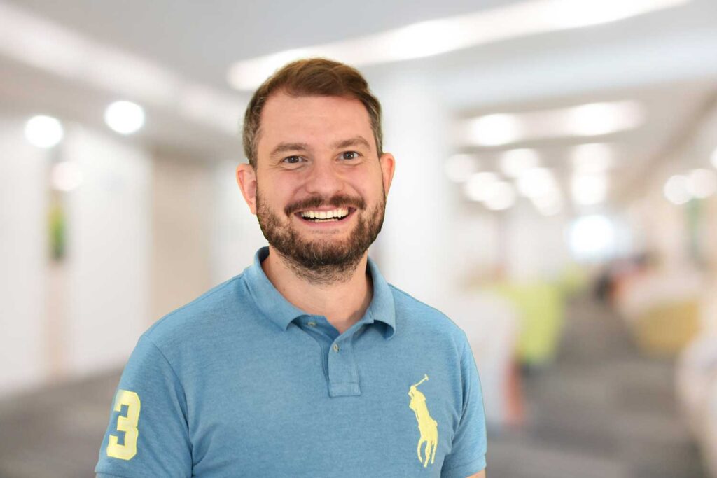 Lächelnder Mann mit Bart und blauem Polohemd, der in einem hellen, unscharfen Innenbereich steht.