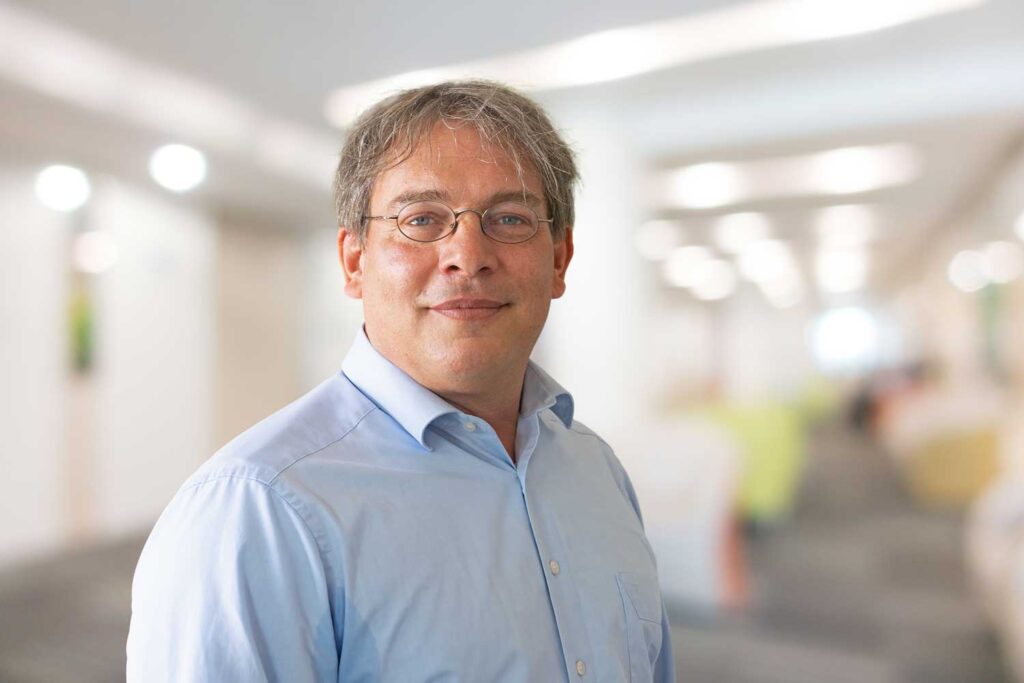 Ein Mann mit Brille und blauem Hemd lächelt in einer hell erleuchteten, verschwommenen Büroumgebung.