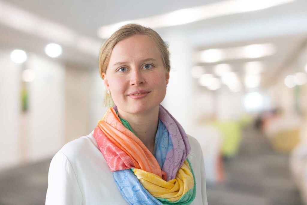 Eine Frau in einem weißen Oberteil mit einem bunten Regenbogenschal steht in einem hellen, verschwommenen Innenraum.