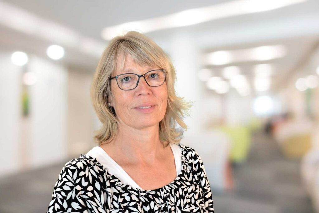 Eine Frau mit Brille und blondem Haar, die ein schwarz-weißes Oberteil trägt, steht in einem hellen, verschwommenen Büro.