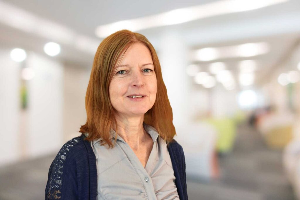 Eine Frau mit rötlich-braunem Haar lächelt in einer hellen, verschwommenen Büroumgebung.