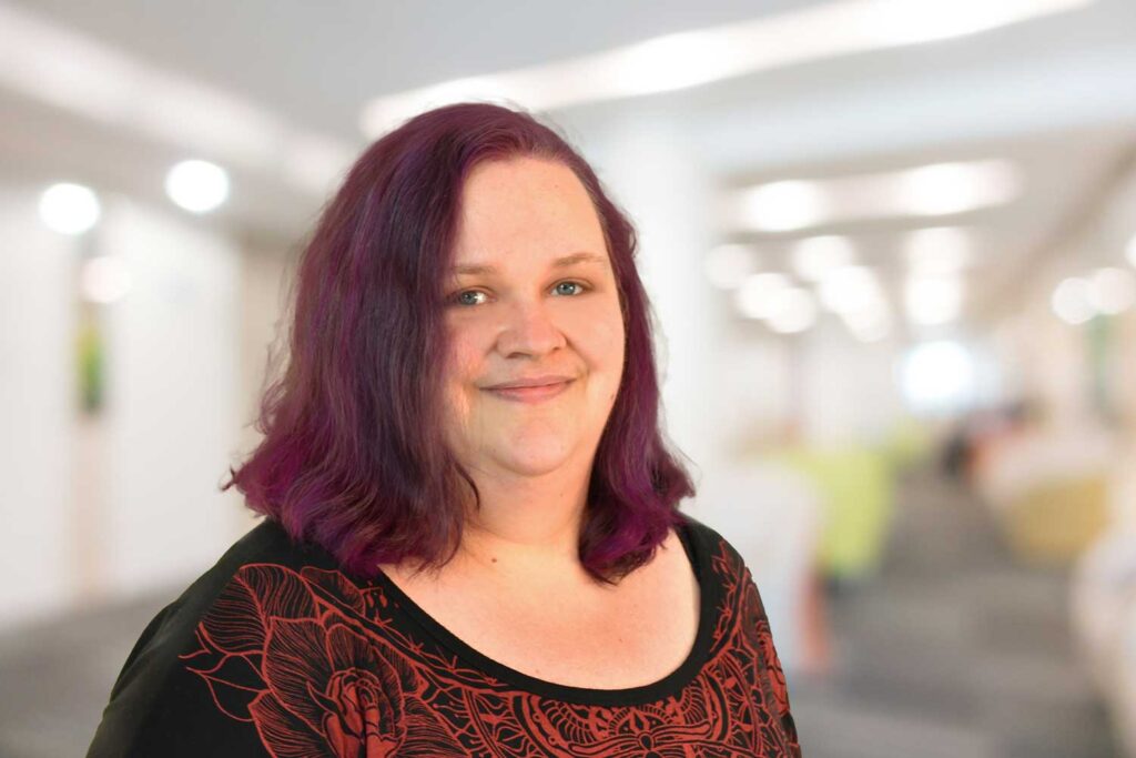 Lächelnde Person mit gewelltem lila Haar in einem schwarz-roten Hemd, die in einem hellen, unscharfen Innenraum steht.