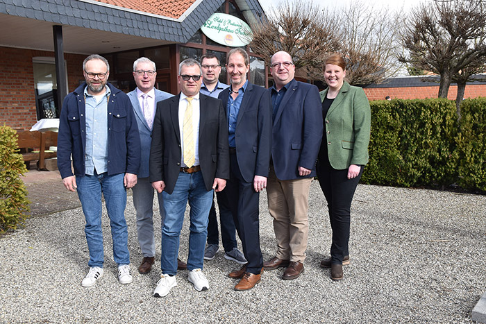 Sieben Erwachsene stehen draußen auf dem Kies vor einem Gebäude und einer Hecke und posieren gemeinsam für ein Gruppenfoto.