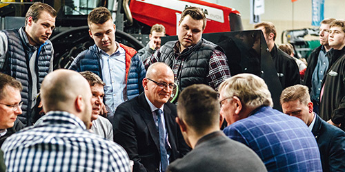 Eine Gruppe diskutierender Männer in Innenräumen, einige mit karierten Hemden und Westen, versammelte sich um die sitzenden Moderatoren.