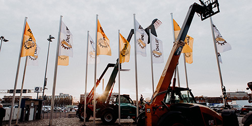 Baufahrzeuge hissen Flaggen in der Nähe von hohen Fahnenmasten mit gelben und weißen Flaggen auf einem Parkplatz bei Sonnenuntergang.