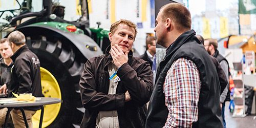Zwei Männer unterhalten sich bei einer Indoor-Veranstaltung vor einem Traktor, während sich andere Personen im Hintergrund unterhalten.