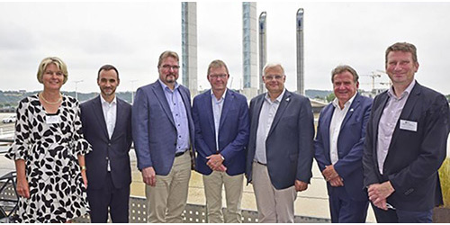 Acht Personen in Geschäftskleidung stehen im Freien mit industriellen Strukturen im Hintergrund.