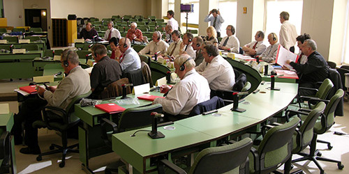 Personen, die in einem Konferenzraum sitzen, Headsets tragen und nach vorne schauen, mit Papieren und Mikrofonen auf den Tischen.