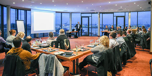 Die Teilnehmer in einem Konferenzraum sehen einem Moderator zu, der eine Präsentation auf einem Bildschirm mit Stadtansichten im Hintergrund hält.