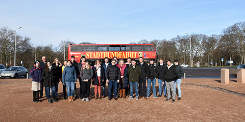 Eine Gruppe von Menschen steht vor einem roten Doppeldeckerbus auf einem Parkplatz.
