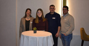Vier Personen, die hinter einem runden Tisch mit einer kleinen Pflanze stehen, posieren und lächeln für ein Foto in einem Innenraum.