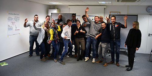 Eine Gruppe von Menschen steht in einem Büro, lächelt und winkt enthusiastisch in die Kamera.