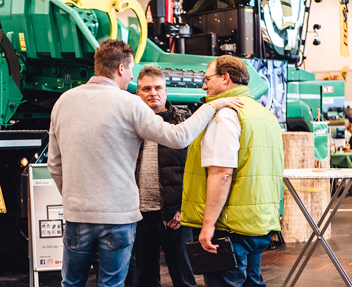 Drei Männer stehen bei einer Indoor-Veranstaltung neben grünen landwirtschaftlichen Geräten und unterhalten sich; einer hat seine Hand auf der Schulter des anderen.