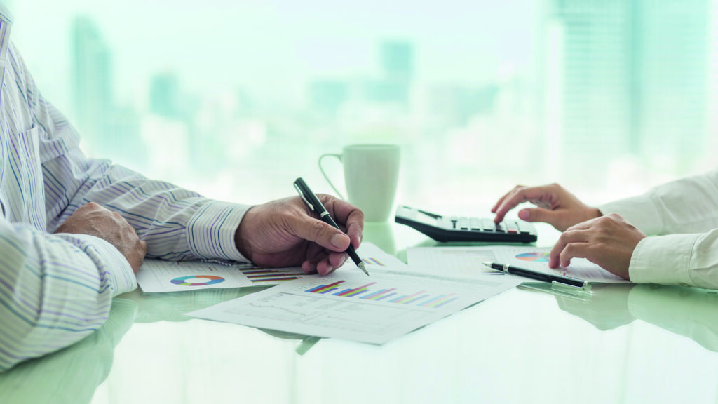 Zwei Personen arbeiten mit Tabellen, Papieren und einem Taschenrechner an einem hellen Bürotisch mit einer Kaffeetasse.